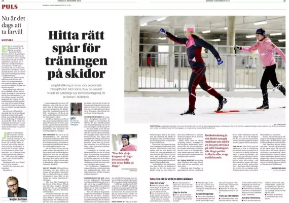 Två personer som åker skidor inomhus på en bana med stavar, bär vintersportutrustning. En person är i rörelse, medan den andra tittar på. Text och artiklar syns i bakgrunden.