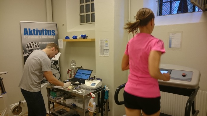 En kvinna joggar på ett löpband iförd en rosa topp, medan en man kontrollerar utrustningen på ett närliggande skrivbord med en bärbar dator. En banderoll för "Aktivitus Testklinik & Coaching" syns i bakgrunden.