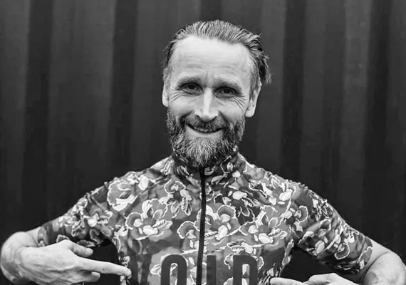 En man med skägg ler och pekar på text på sin blommönstrade cykeltröja. Svartvitt foto.