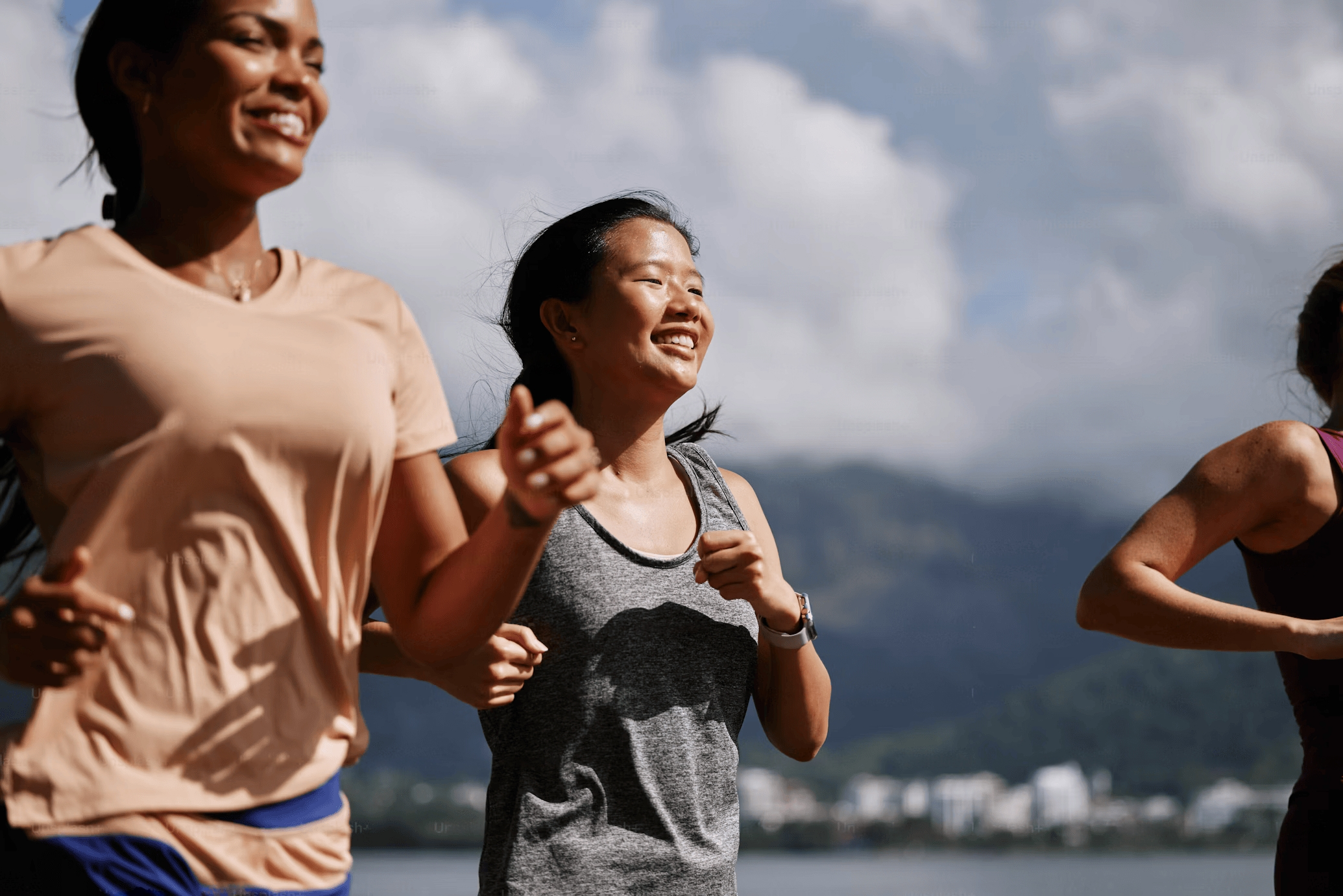 Tre personer joggar utomhus nära vattnet med berg och moln i bakgrunden.