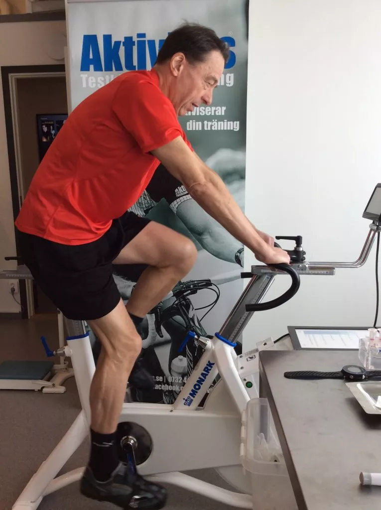 Man i röd skjorta och svarta shorts pedaler på en stationär cykel inomhus, med en banderoll i bakgrunden och ett bord i närheten.