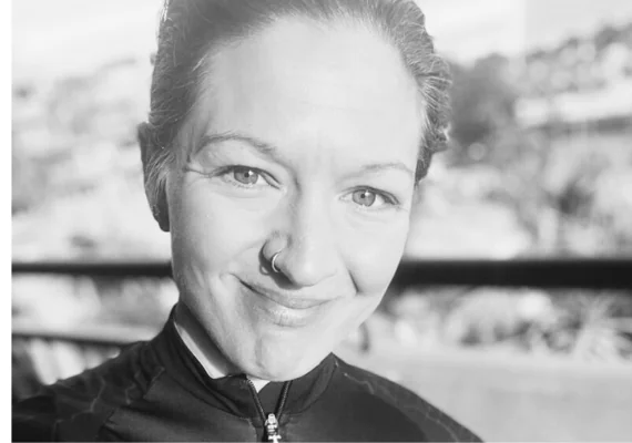 En person med näsring och kort hår ler i ett svartvitt närbildsporträtt, med suddig utomhusbakgrund.