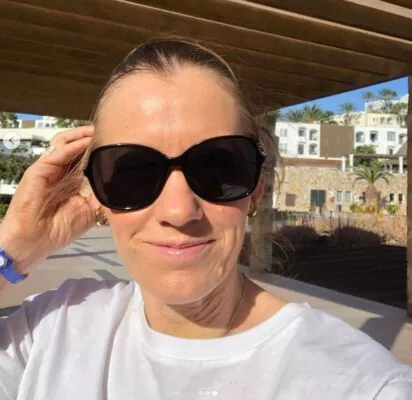 Person som bär solglasögon och en vit topp tar en selfie utomhus med byggnader i bakgrunden.