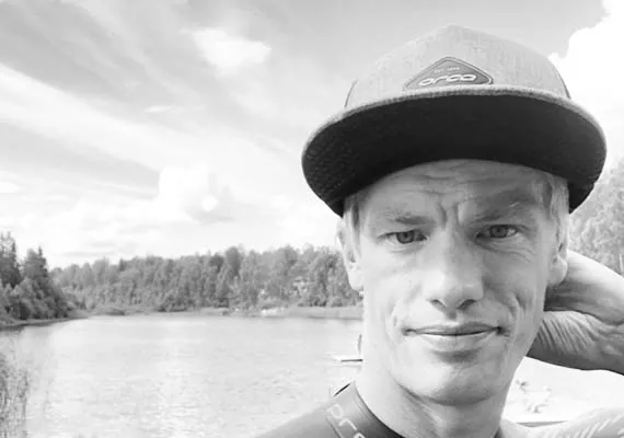 Man i keps och fritidskläder står vid en flod med träd i bakgrunden en klar dag.