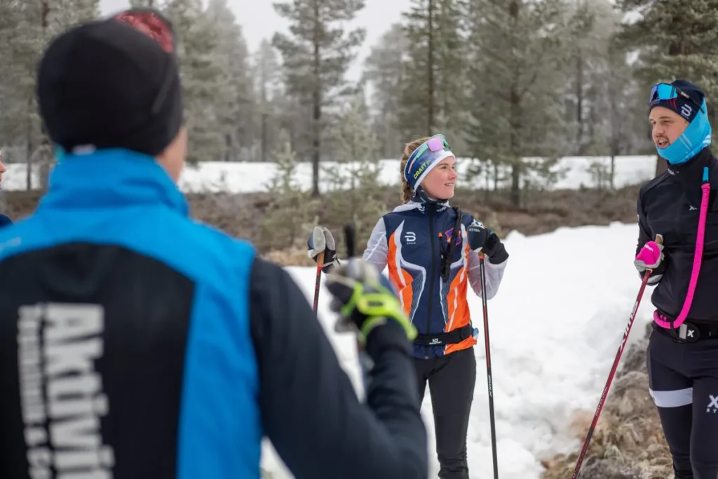 Tre personer i vintersportkläder står på snötäckt mark i en skog, håller i skidstavar och bär hattar och jackor.