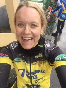 En leende cyklist med ett lerigt ansikte iklädd en gul och svart tröja står utomhus. Ytterligare en cyklist står i bakgrunden.