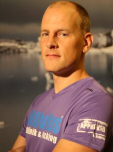 En person i en lila skjorta står med armarna i kors mot en suddig bakgrund av vatten och stenar. - Aktivitus En person i en lila skjorta står med armarna i kors mot en suddig bakgrund av vatten och stenar.