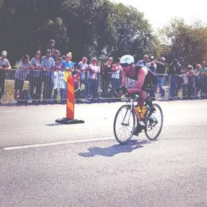 Cyklist i ett lopp med hjälm och solglasögon cyklar på en solig väg, med åskådare bakom en barriär. - Aktivitus Cyklist i ett lopp med hjälm och solglasögon cyklar på en solig väg, med åskådare bakom en barriär.