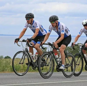 Tre cyklister i matchande tröjor och hjälmar cyklar på en väg med grönska och ett avlägset vatten i bakgrunden. - Aktivitus Tre cyklister i matchande tröjor och hjälmar cyklar på en väg med grönska och ett avlägset vatten i bakgrunden.