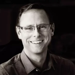 En person ler medan han bär glasögon och en mönstrad skjorta, med en mörk bakgrund. - Aktivitus En person ler medan han bär glasögon och en mönstrad skjorta, med en mörk bakgrund.