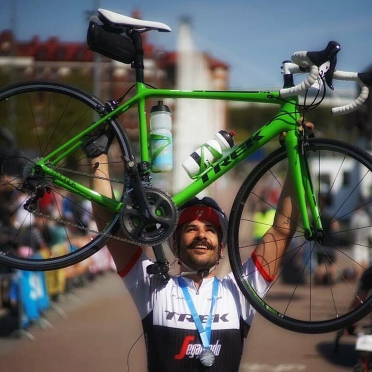 Cyklist i racerutrustning håller triumferande en grön landsvägscykel över huvudet vid ett utomhusevenemang, omgiven av byggnader och åskådare.