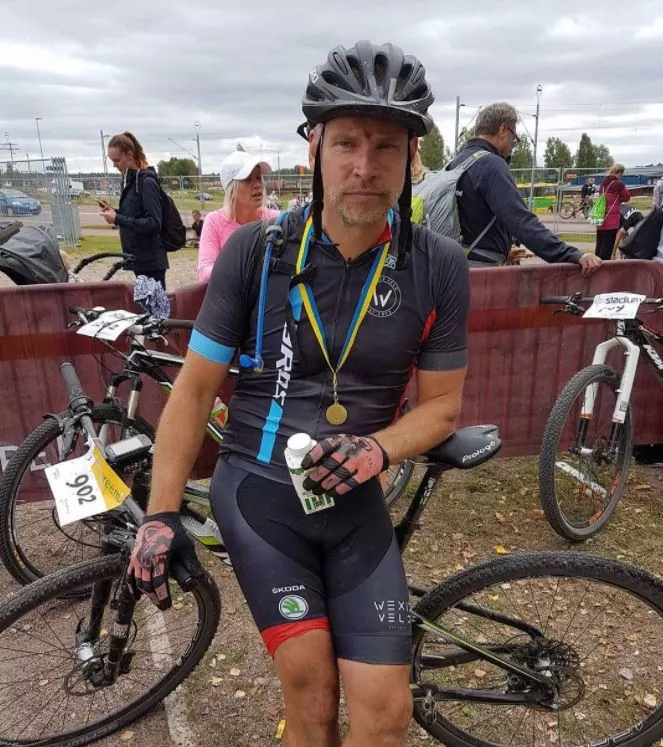 Cyklist i racerutrustning håller en drink, bär hjälm och medaljer, lutad på en mountainbike. Andra cyklister i bakgrunden, med mulen himmel ovanför.