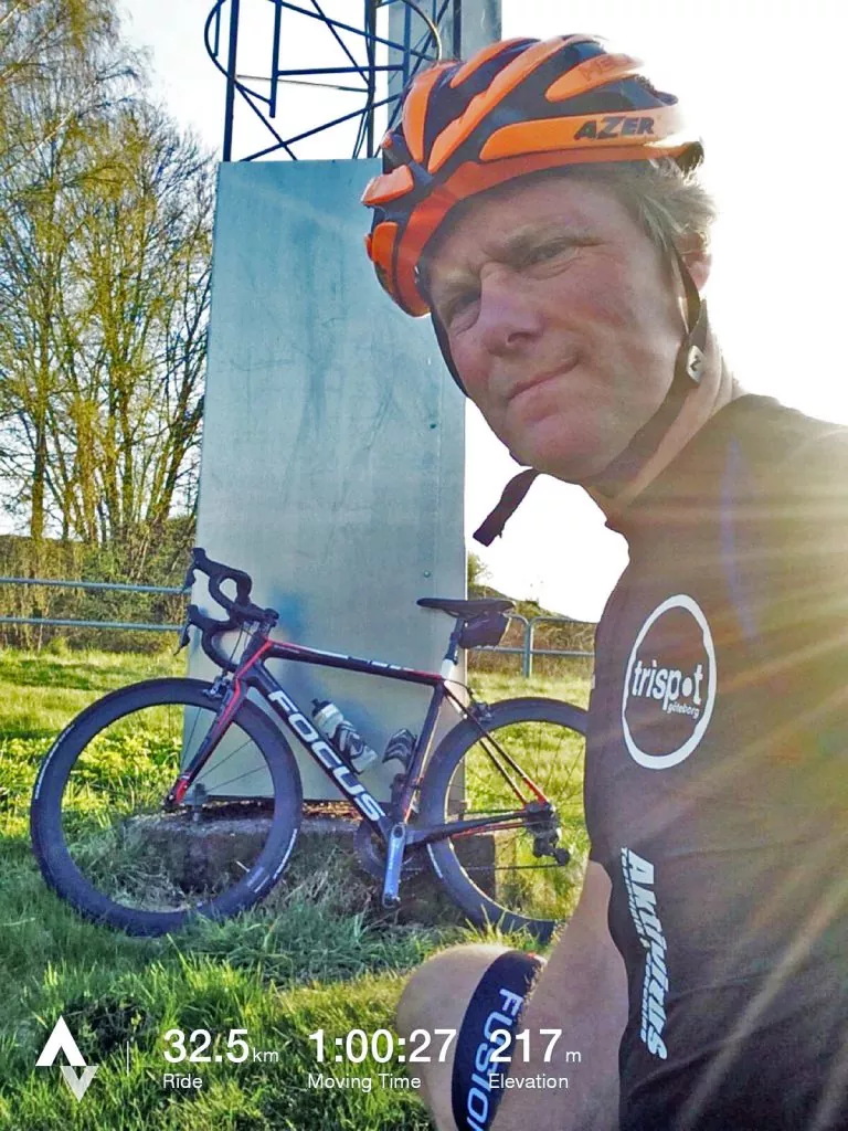 En cyklist som bär en orange hjälm tar en selfie bredvid en cykel. Bilden visar åkdetaljer: 32,5 km distans, 1:00:27 tid, 217 m höjd. Solljus och en metallstruktur är synliga.