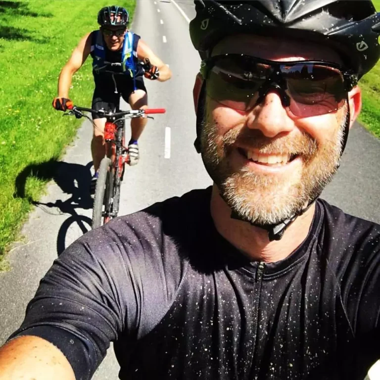 Två cyklister som bär hjälmar och solglasögon cyklar på en solig väg. Förgrundscyklisten tar en selfie med den andra cyklisten bakom, med gräset synligt till vänster.