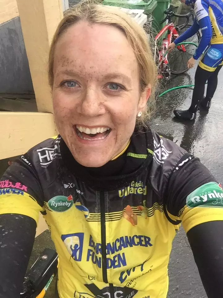 En leende cyklist med ett lerigt ansikte iklädd en gul och svart tröja står utomhus. Ytterligare en cyklist står i bakgrunden.