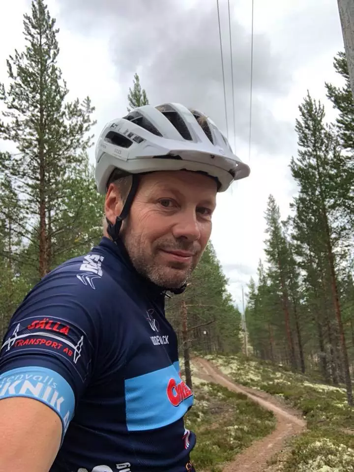 En man i cykelhjälm och tröja står på en skogsstig, omgiven av höga träd och en molnig himmel.