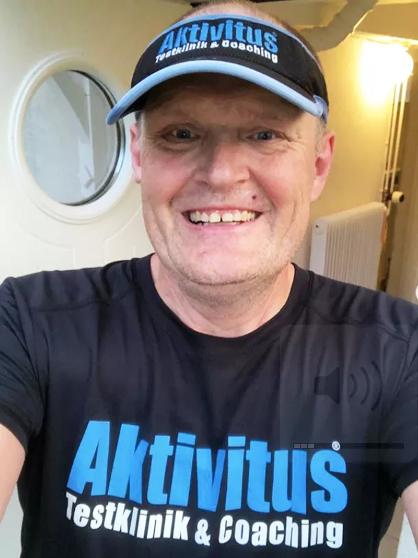Man leende, klädd i svart och blå Aktivitus Testklinik & Coaching-keps och skjorta, framför ett runt fönster.