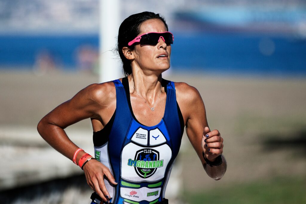 En person som springer utomhus, iklädd blå triathlondräkt och rosa solglasögon, med strand och suddig bakgrund.