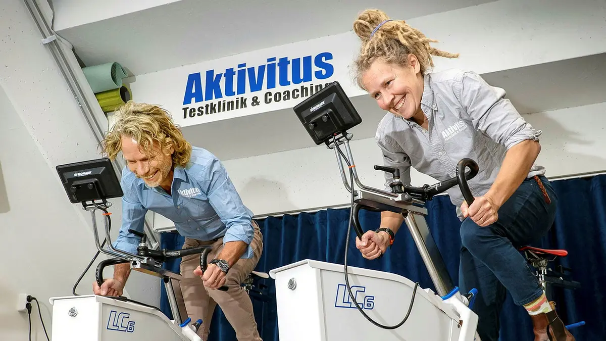 Två leende personer cyklar stillastående inomhus, iklädda matchande Aktivitus-skjortor. På en skylt ovan står det "Aktivitus Testklinik & Coaching.