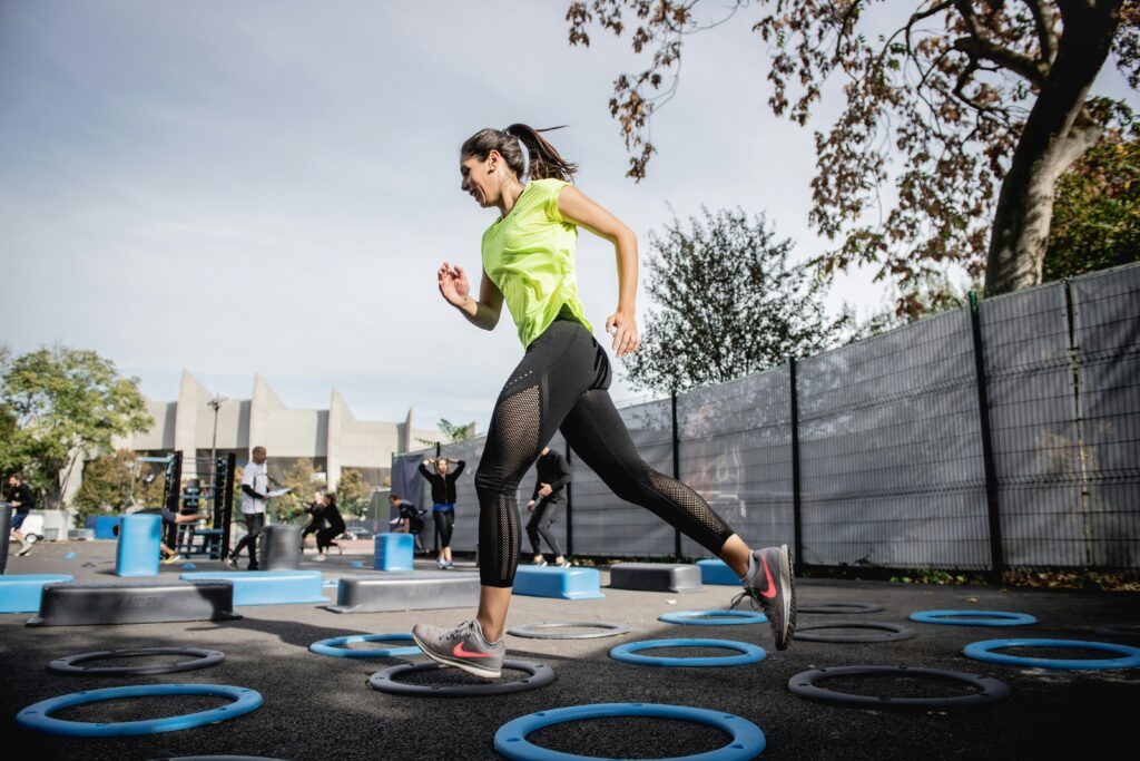 En kvinna i en ljusgrön skjorta och svarta leggings hoppar över blå runda plattformar på en utomhus fitnesspark. Andra tränar i bakgrunden.