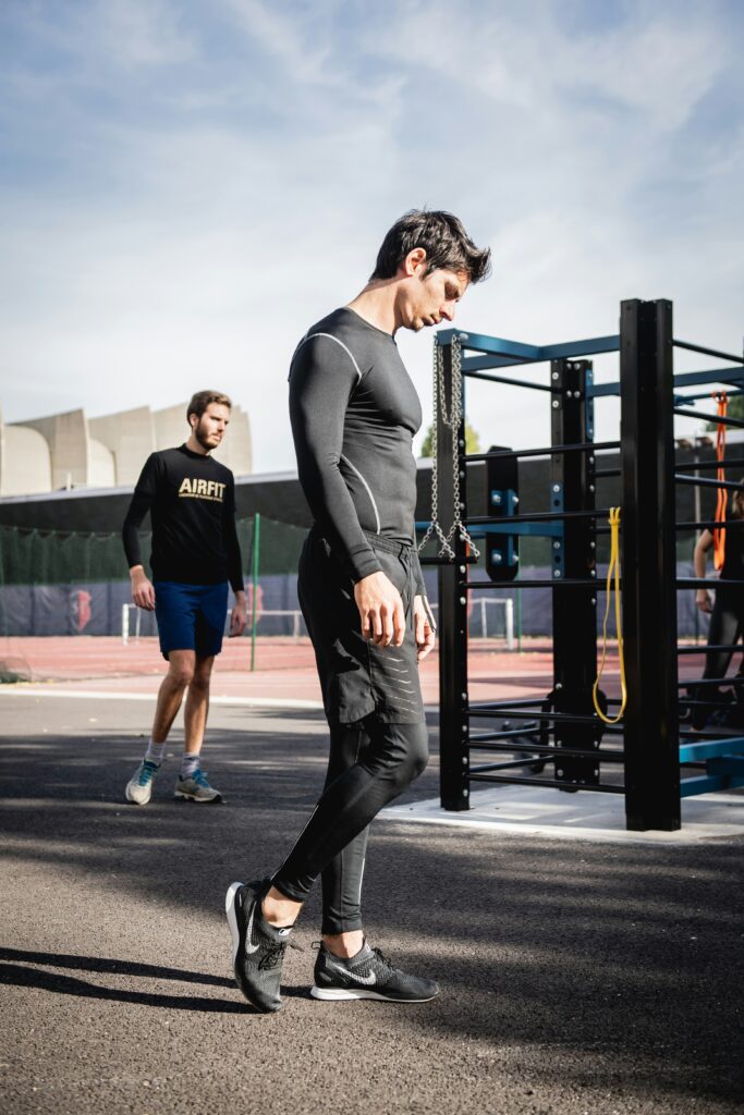 Två män i sportkläder är på ett utegym. Den ena går medan den andra, i bakgrunden, joggar. Träningsutrustning är synlig nära dem under en klar himmel.
