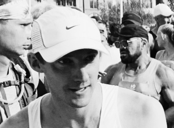 Runner in a white Nike cap at the start of a crowded race, smiling slightly as others prepare behind him.