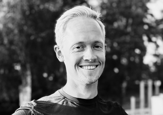 Smiling man with short blond hair wearing a dark shirt outdoors, blurred trees in the background (black and white).