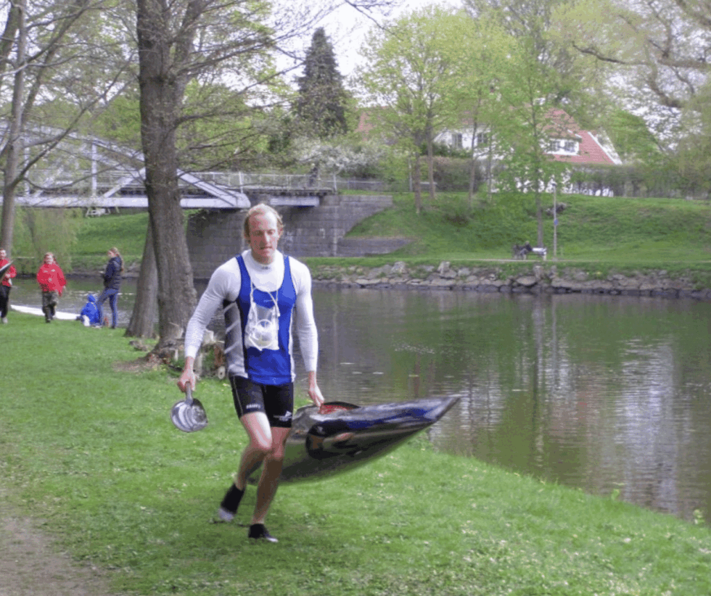 Man in a blue and white jersey carrying a small kayak along a grassy lakeside path, trees and a bridge in the background