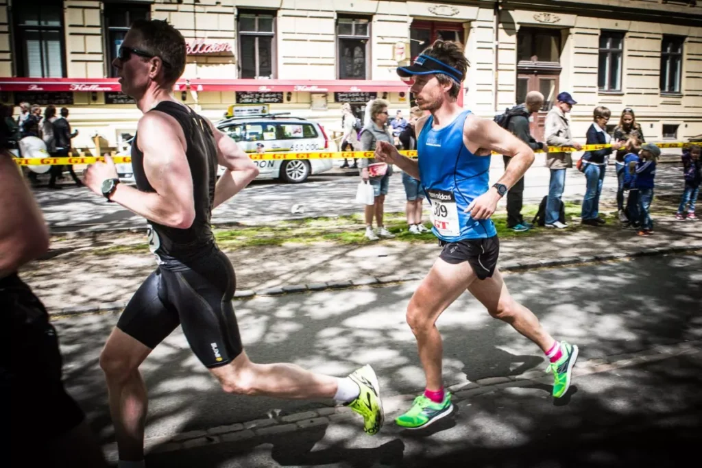 Två löpare i atletisk utrustning deltar i ett lopp, med åskådare som tittar på från trottoaren. De går förbi en byggnad klädd med försiktighetstejp.