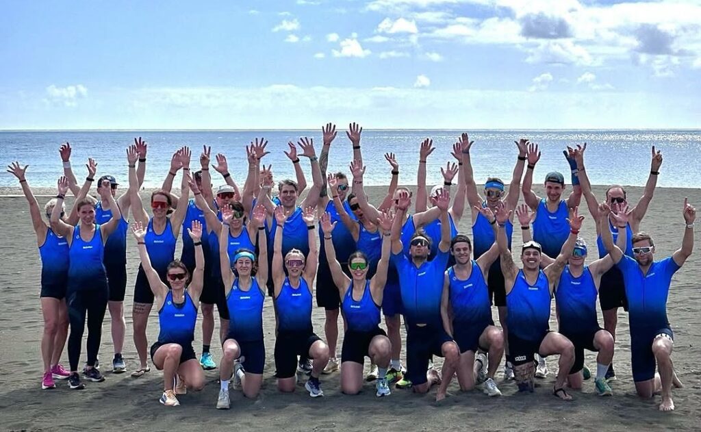 Grupp människor som bär blå toppar och svarta underdelar, poserar med upphöjda armar på en strand under en delvis molnig himmel.