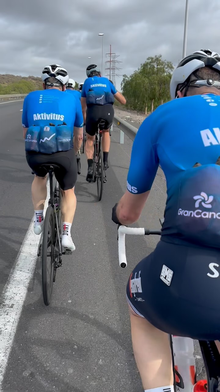 Tre cyklister i blå Aktivitus-tröjor cyklar på en väg, med molnig himmel ovanför.