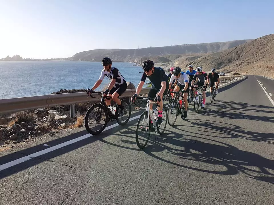 Cyklister cyklar i rad längs en väg vid havet under en klarblå himmel, med kullar i bakgrunden och solljus som kastar skuggor.