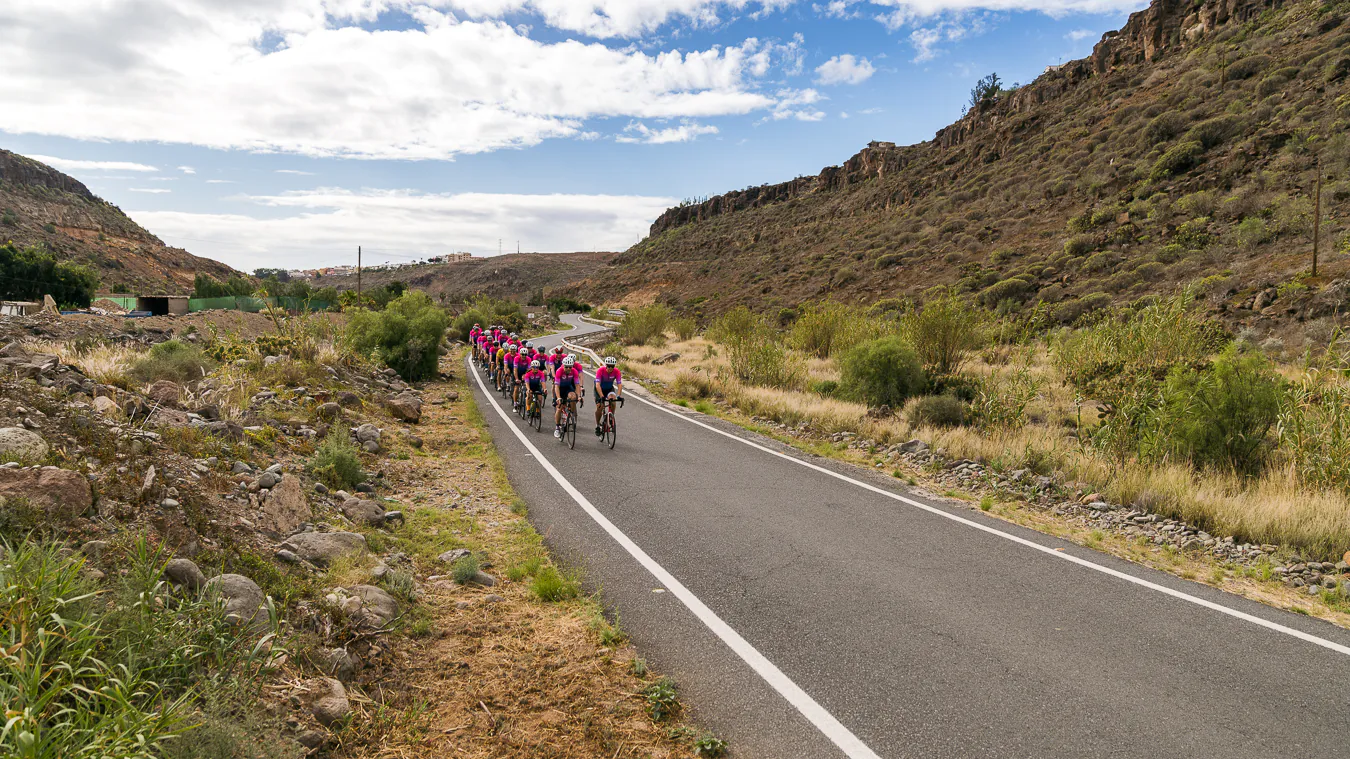 En grupp cyklister i rosa tröjor cyklar på en asfalterad väg genom ett stenigt landskap under en halvmolnig himmel.