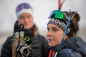 Young woman in a winter jacket with mirrored ski goggles on her head, holding climbing gear; a teammate is blurred in the background.