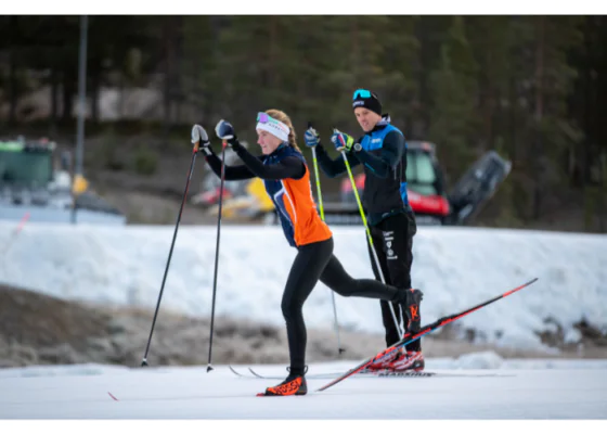- Aktivitus Two cross-country skiers racing on a snowy track; woman in an orange vest leads, man in blue/black follows, with trees and vehicles in the background.