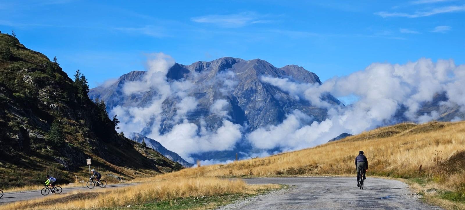 Cyclists ride along a winding mountain road with golden grass and rugged peaks under a blue sky with clouds in the distance.