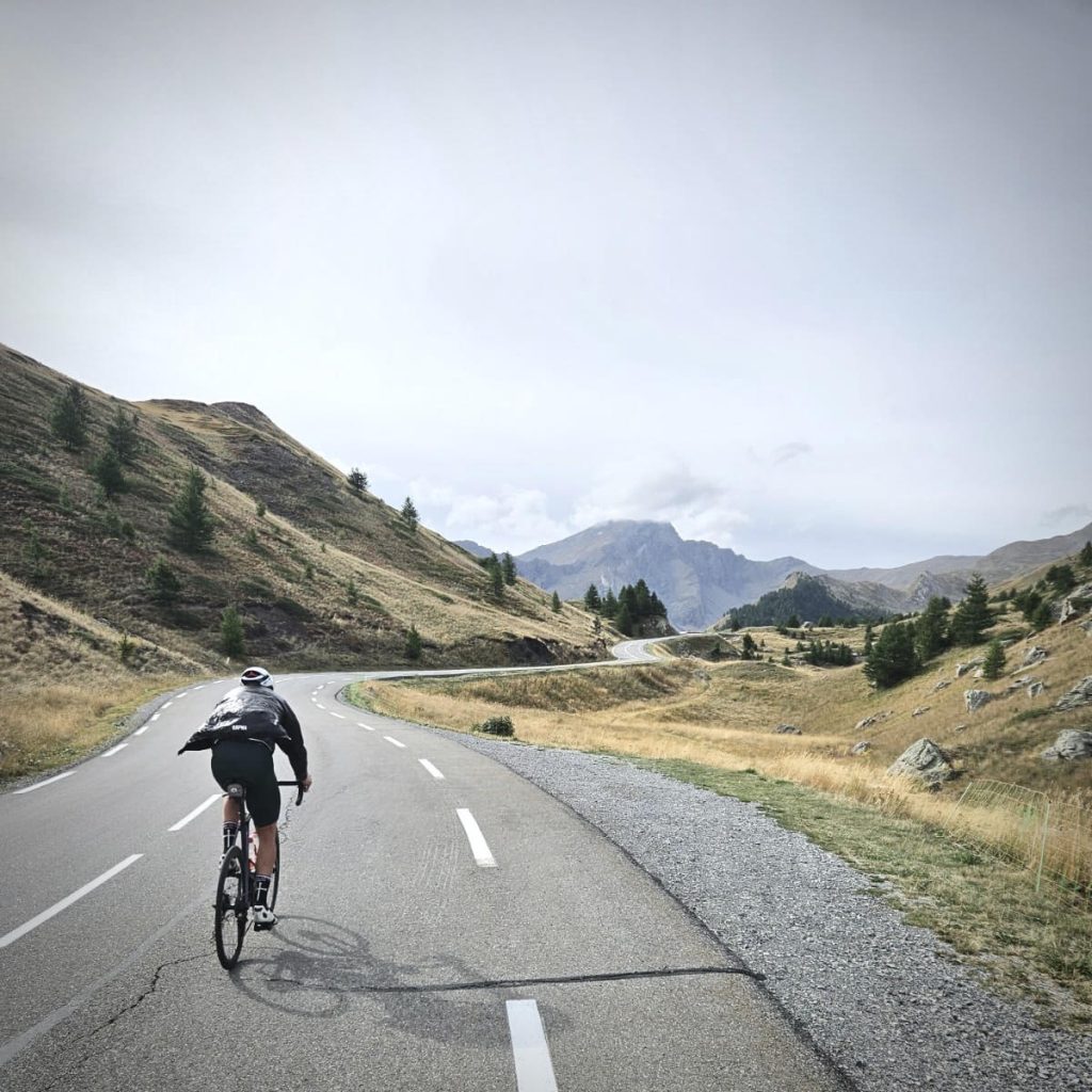 Cyclist wearing a helmet rides a road bike on a winding mountain road with grassy hills and distant mountains under a cloudy sky, ahead in the distance.