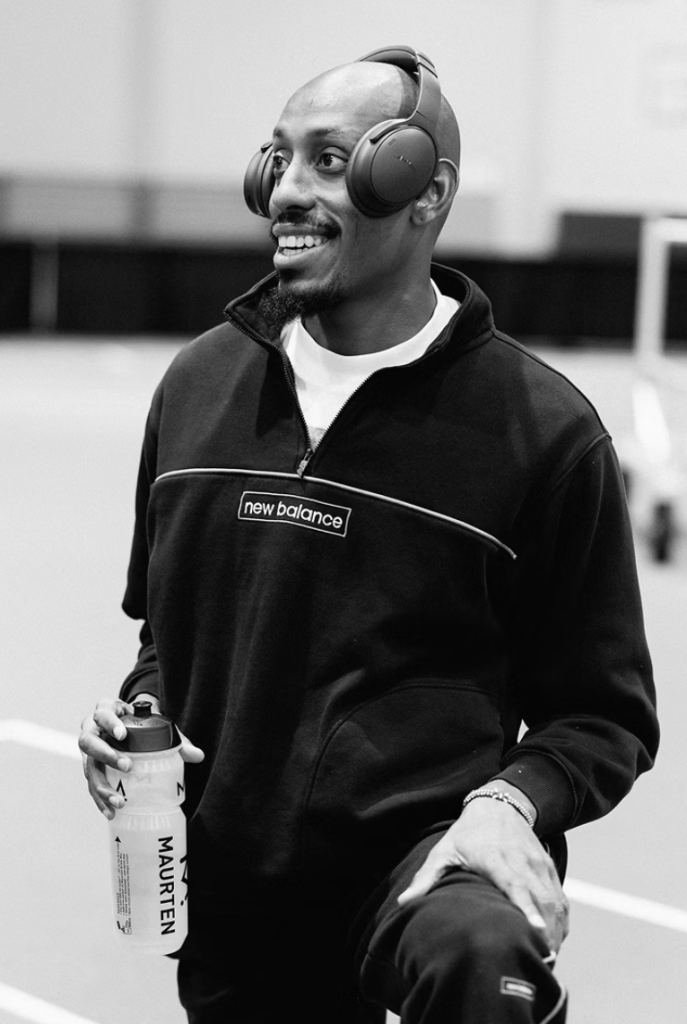 Skärmavbild 2026-05-08 kl 083355 - Aktivitus Smiling man in a black New Balance pullover wearing large over-ear headphones, holding a Maurten water bottle in a gym (black-and-white photo).