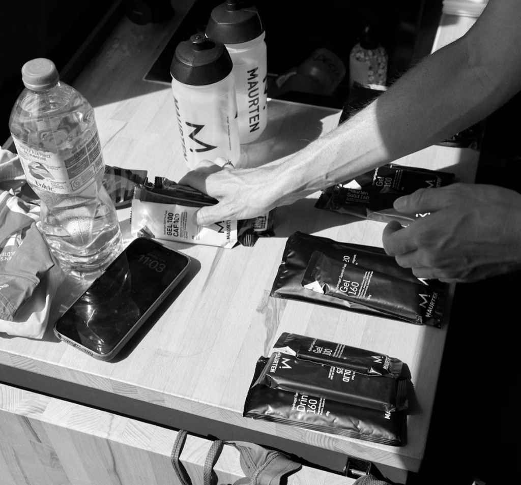 Skärmavbild 2026-05-08 kl 083453 - Aktivitus Hands grabbing a Maurten energy gel packet on a wooden table with water bottles and a smartphone nearby.