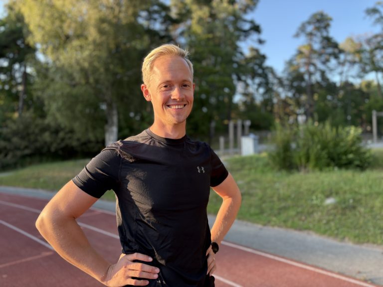 Smiling man in a black athletic shirt standing on an outdoor running track with hands on hips.