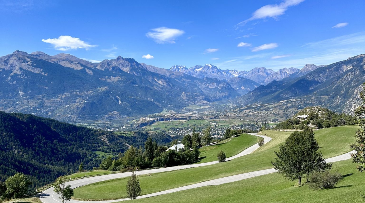 Wide alpine valley with winding road, green fields, and distant snow-capped mountains under a bright blue sky.