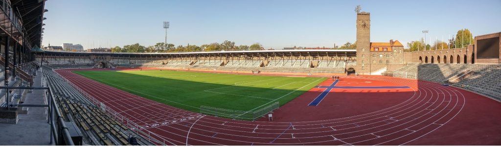 Namnlös design 4 - Aktivitus Panoramic view of an outdoor stadium with a red running track surrounding a green field and brick stands on both sides under a clear blue sky