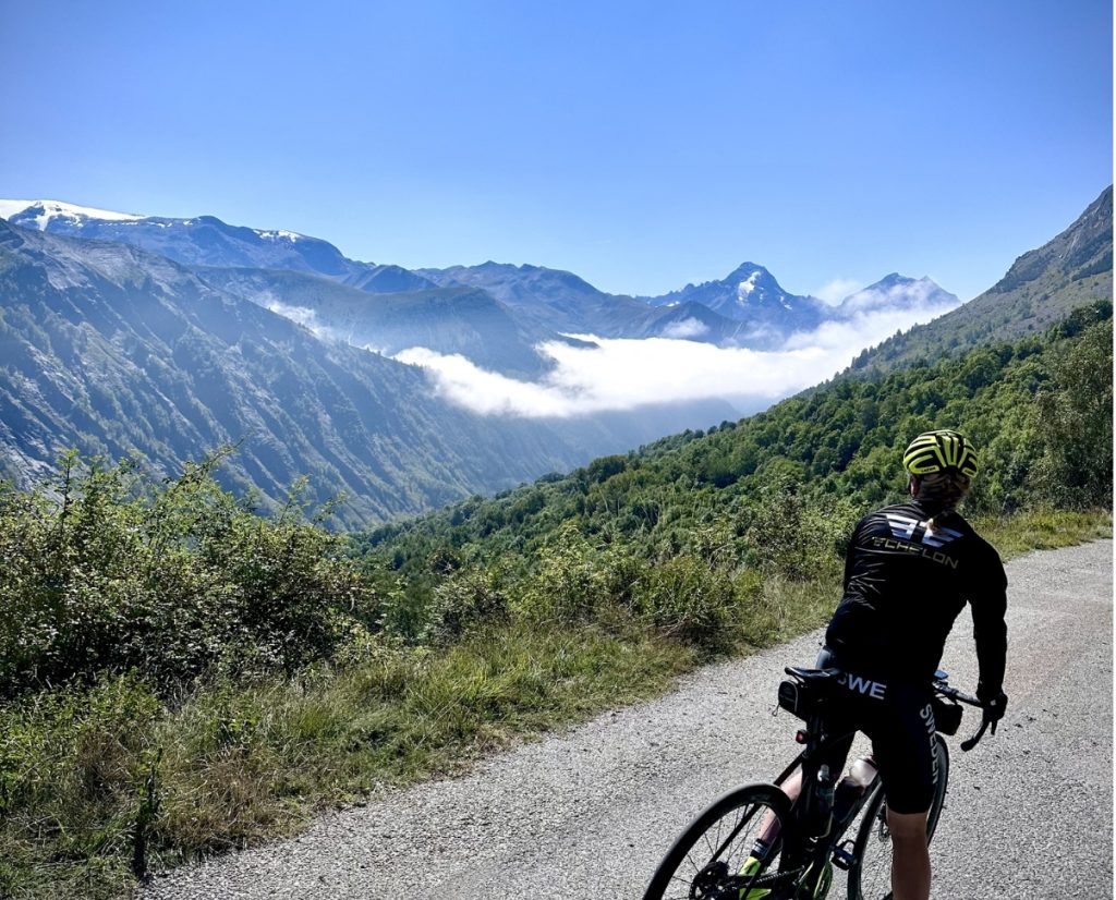 Cyclist in helmet riding a mountain road with a valley, snow-capped peaks, and a clear blue sky in the background