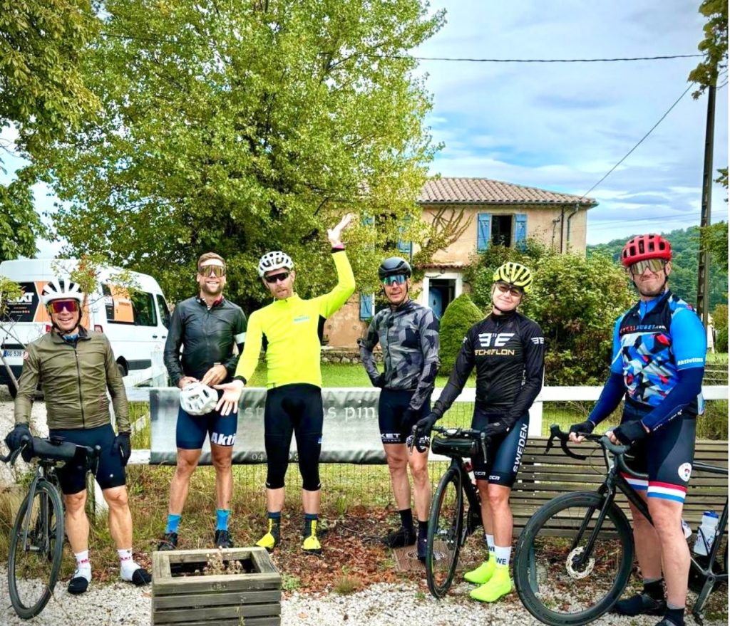 Group of cyclists posing with bikes outdoors in front of a house, wearing helmets and colorful cycling gear.