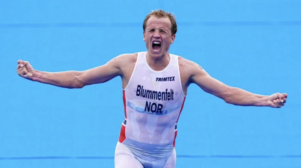 Male athlete in a white Norwegian racing suit, arms outstretched in celebration against a blue background.