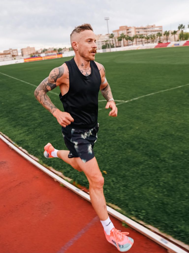 Tattooed man in a black sleeveless shirt sprinting on a red track by a green field with buildings in the background.