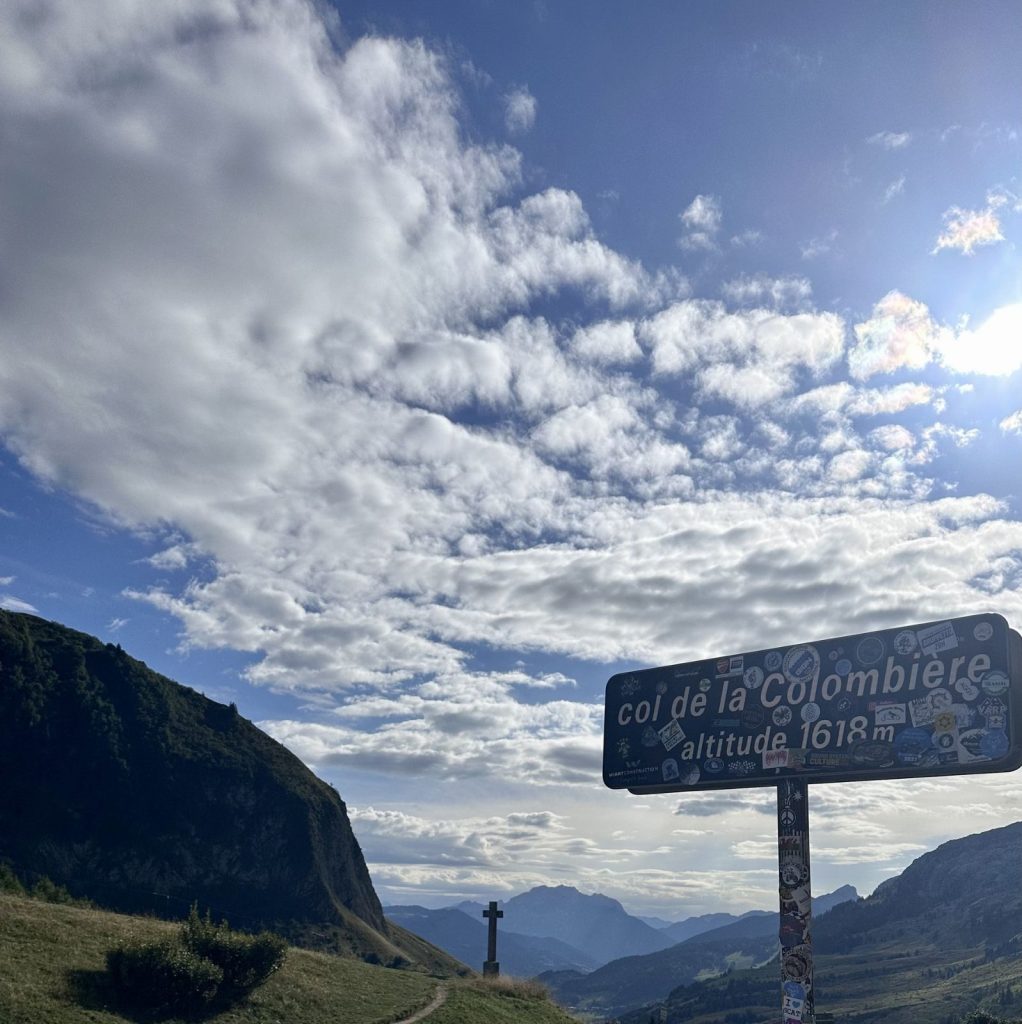 Signpost in a mountain valley showing Col de la Colombière, altitude 1618 m, with stickers on the sign.