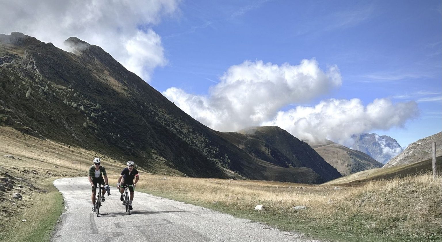 Two cyclists ride along a mountain road with rocky slopes and blue sky above.