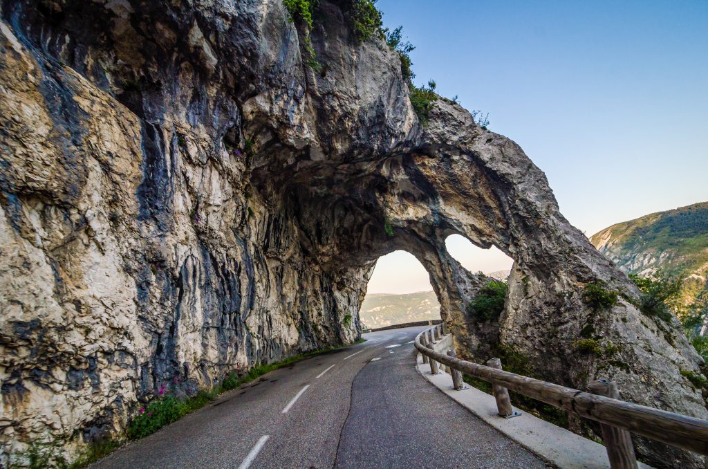 Winding mountain road passes beneath a natural rock arch, with a guardrail and valley below? Actually, without question mark.