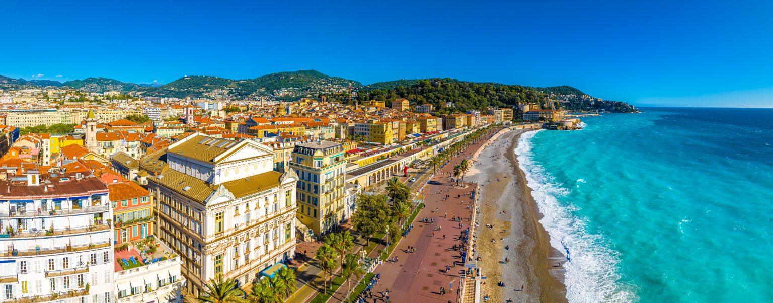 Panoramic aerial view of a sunny Mediterranean city along a palm-lined promenade, colorful buildings, and a turquoise sea.